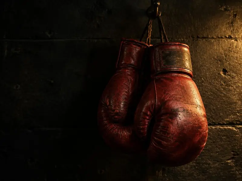 A worn pair of red leather boxing gloves hanging from a hook on a dark wall